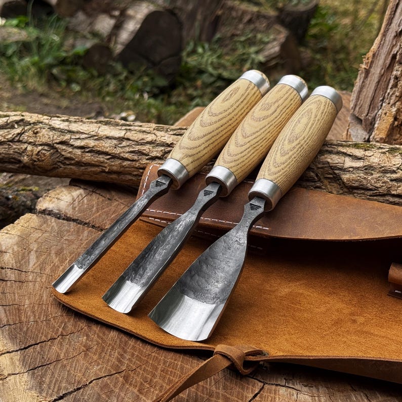 May include: Three wood carving chisels with light-coloured wooden handles and silver metal ferrules. The blades are made of dark, forged steel with varying widths, including a narrow gouge, a medium gouge, and a wide, flat-edged chisel blade.