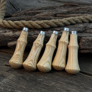 以下が含まれることがあります： 木目模様の明るい木材で作られた5つのハンドルと、銀色の金属キャップ。 ハンドルは、風化した木材の表面に一列に並んでいます。 太いロープが背景に見えます。