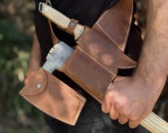 May include: A brown leather axe sheath with a matching shoulder strap. The sheath holds a silver axe head with a wooden handle. The leather is stitched with white thread and has a snap closure.