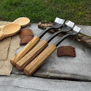 May include: Four wood carving gouges with wooden handles and curved metal blades. The tools are arranged on a weathered wooden surface, with a wooden spoon, leather sheaths, and a piece of bark.