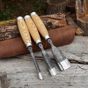 May include: Three woodworking chisels with light-coloured wooden handles and silver metal ferrules. The steel blades feature a dark, forged finish with a polished cutting edge. Each tool has a distinct blade width, ranging from narrow to wide.
