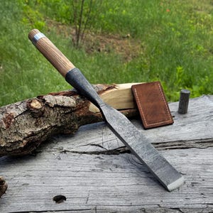 May include: A hand-forged steel wood chisel featuring a long, textured metal blade and a cylindrical wooden handle with a silver-colored metal ferrule. The tool is positioned alongside a small, rectangular brown leather sheath and a wooden log.