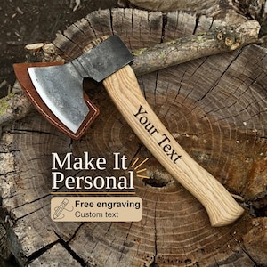 Peut inclure: Une hachette avec une tête en métal forgé sombre, un manche en bois brun clair et un étui en cuir brun. Le manche porte le texte gravé "Your Text". L'image contient le texte "Make It Personal" et "Free engraving Custom text".