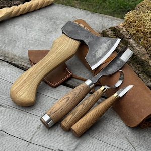 May include: A collection of wood carving tools, including a small axe with a wooden handle, and three carving knives with wooden handles and metal blades. The tools are displayed on a weathered wooden surface, with leather sheaths.