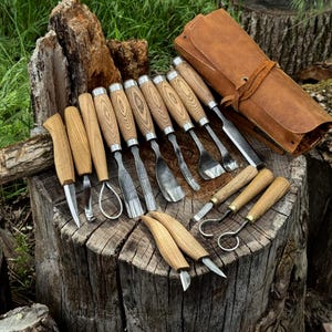 May include: A collection of wood carving tools with wooden handles and metal blades, arranged on a tree stump. The set includes various gouges, knives, and a leather tool roll. The tools are designed for woodworking and carving projects.