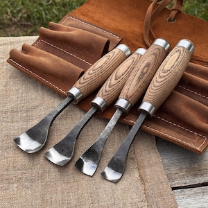 May include: Four wood carving gouges with wooden handles and metal blades are displayed next to a brown leather tool roll. The gouges have a curved blade design and are made of metal and wood.