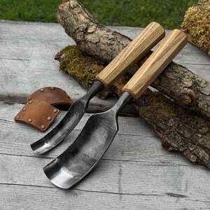 May include: Two wood carving gouges with wooden handles and curved steel blades. The tools are displayed on a weathered wooden surface, alongside a small leather sheath with rivets and pieces of wood.