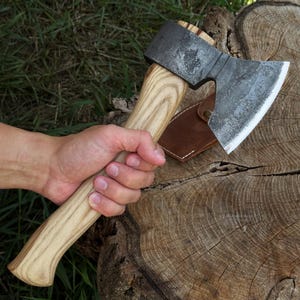 May include: A hand axe with a forged, dark grey metal head featuring a sharpened silver edge and a light-colored, natural wood grain handle. A brown leather sheath with a metal snap button is positioned near the axe head on a wooden surface.