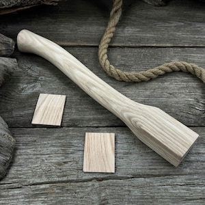 May include: A light-colored wooden axe handle with a slightly curved shape. The handle is lying on a wooden surface with two small wooden blocks next to it.