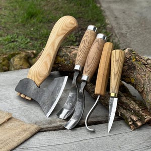 May include: A collection of woodworking tools, including a small axe with a light wood handle and a dark metal head, along with several carving knives and gouges. The tools are arranged on a wooden surface, showcasing their craftsmanship.