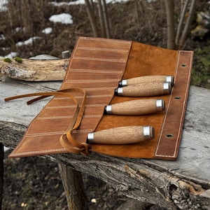 May include: A brown leather tool roll featuring multiple stitched slots and a long leather tie closure. Three wooden-handled tools with silver metal ferrules are inserted into the slots, showcasing a natural grain texture and cylindrical shape.