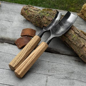 May include: Two curved wood carving gouges with wooden handles and metal blades. The gouges are positioned diagonally on a wooden surface, with two brown leather sheaths and a piece of bark in the background. The gouges are designed for woodworking.