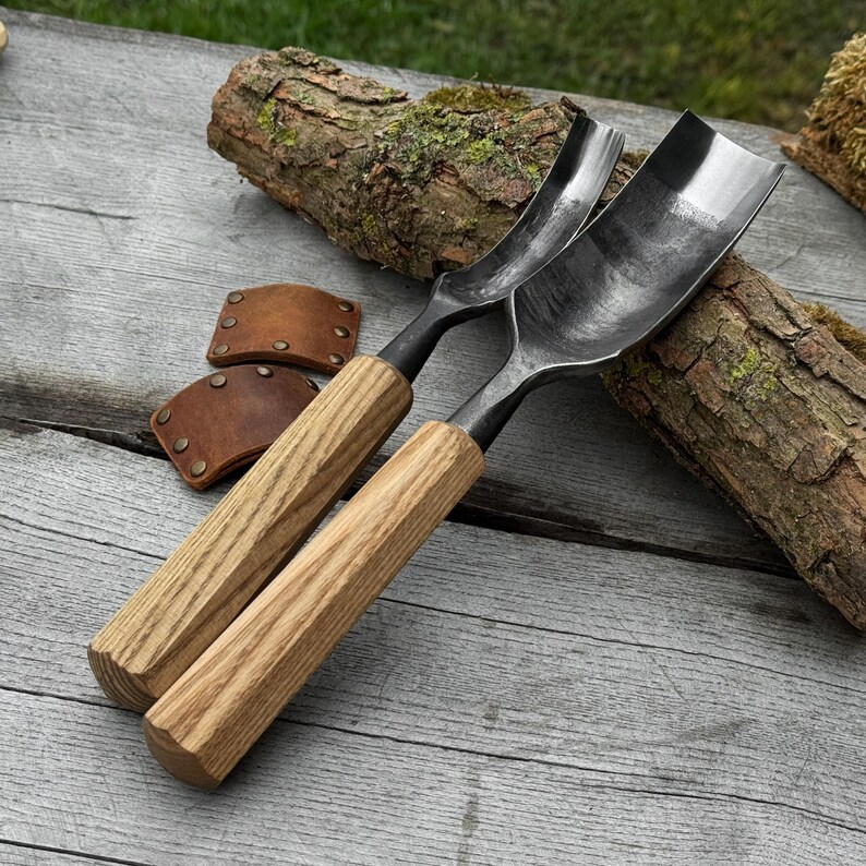 May include: Two curved wood carving gouges with wooden handles and metal blades. The gouges are positioned diagonally on a wooden surface, with two brown leather sheaths and a piece of bark in the background. The gouges are designed for woodworking.