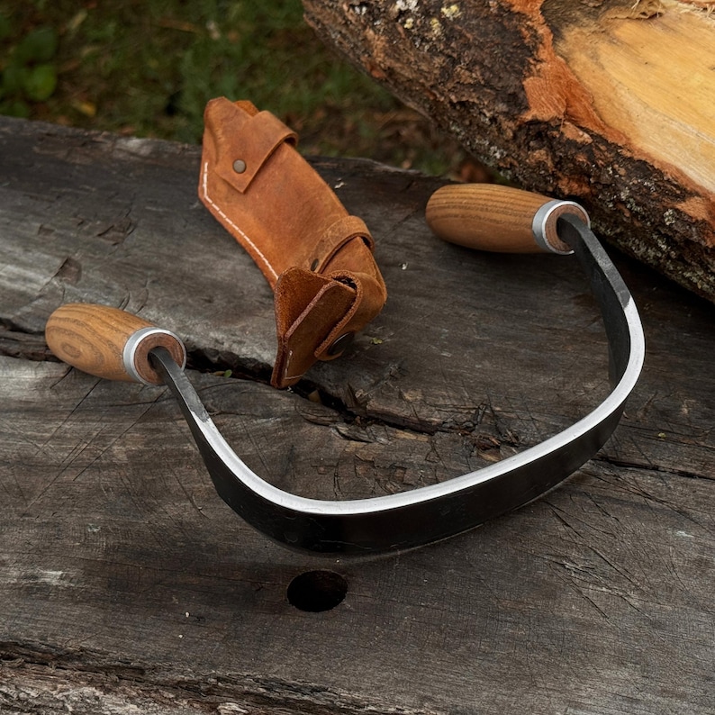 May include: A curved draw knife with wooden handles and a leather sheath. The blade is made of steel and has a sharp edge. The handles are made of wood and are attached to the blade with metal rings. The sheath is brown and protects the blade.