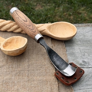 May include: A wood carving spoon gouge with a wooden handle and a metal blade. The gouge is resting on a brown leather sheath. Two carved wooden spoons are in the background.