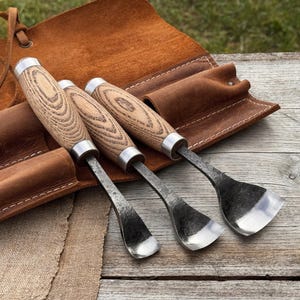 May include: Three wood carving chisels with wooden handles and metal blades, resting on a brown leather tool roll. The chisels have a silver metal band where the handle meets the blade. The wood grain is visible on the handles.