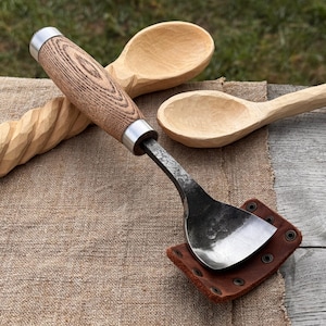 May include: A handcrafted spoon carving tool with a wooden handle and a metal blade, resting on a leather sheath. Two wooden spoons are in the background, along with a carved wooden piece. The tools are on a burlap surface.