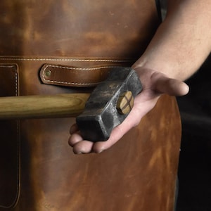 May include: A blacksmith's hammer with a wooden handle and a dark, textured metal head. The hammer's head has a decorative wooden accent. The background includes a leather apron. The hammer is a tool for shaping metal.