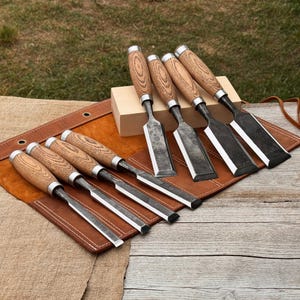 May include: A set of seven woodworking chisels with light brown wooden handles and dark metal blades. Four chisels are arranged in a row on a brown leather tool roll, while three chisels are positioned upright against a small rectangular wooden block.