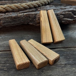Op de afbeelding: Zes onafgewerkte houten blokken met een natuurlijk houtnerfpatroon. De blokken zijn rechthoekig met schuine zijden en platte uiteinden. Ze zijn gerangschikt op een verweerd houten oppervlak.