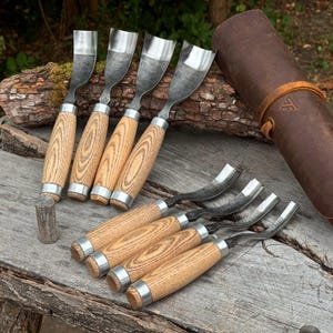 May include: A set of eight wood carving gouges with curved metal blades and light-colored wooden handles. Four gouges are arranged vertically with wide, curved blades, while four are positioned horizontally. A brown leather tool roll is included.