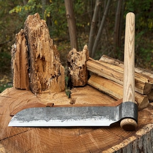 May include: A hand-forged, heavy-duty hatchet with a wooden handle and a wide, textured blade. The hatchet is resting on a tree stump, with a leather sheath visible. The background includes firewood and a natural forest setting.