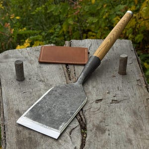 Puede incluir: Un cincel de metal grande, forjado a mano, con mango de madera. El cincel tiene una hoja ancha y rectangular con un borde afilado. También se ve una pequeña cartera de cuero marrón y dos clavijas de madera.