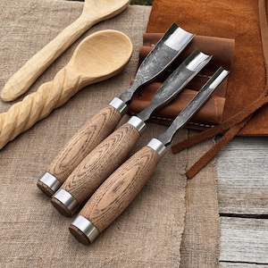 Puede incluir: Un conjunto de herramientas para tallar madera, que incluye tres gubias con mangos de madera y hojas de metal. También se ven dos cucharas de madera, una con mango retorcido y otra con cuenco redondeado. Las herramientas descansan sobre una superficie texturizada de tono neutro.