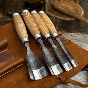 May include: A set of four woodworking chisels with light brown wood handles and silver metal ferrules. The steel blades feature a dark, textured finish with polished, sharp edges. The tools are arranged side by side on a brown leather roll.