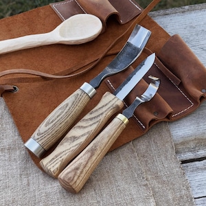 May include: A set of wood carving tools with wooden handles and metal blades, including a spoon carving tool, resting on a brown leather tool roll. The tools have a natural wood grain finish.