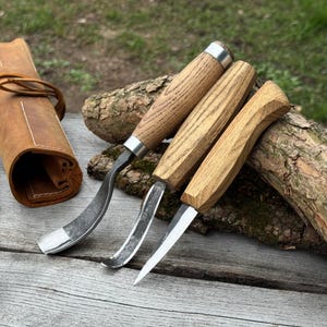 May include: Three wood carving tools with curved and angled blades, featuring light-colored wooden handles and silver metal accents. A rolled-up brown leather tool roll is visible on the left. The tools are arranged on a weathered wooden surface.
