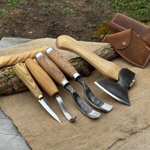 Könnte beinhalten: Eine Sammlung von Holzschnitzwerkzeugen, darunter Hohleisen, ein Messer und eine kleine Axt, mit Holzgriffen und Metallklingen. Die Werkzeuge sind auf einer beigen Jutefläche angeordnet, mit einer Lederscheide und einem geschnitzten Holzstück.