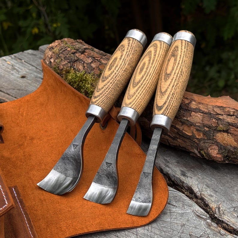 May include: Three wood carving tools with curved, dark metal blades and light-colored, textured wooden handles. Each tool features a silver-toned metal ferrule connecting the blade to the handle and a small triangular maker's mark on the metal.