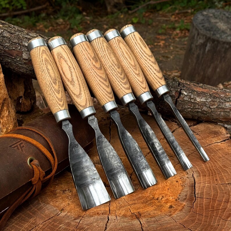 May include: A set of six hand-forged steel wood carving chisels with light-colored wooden handles and metal ferrules, arranged in a row. The tools feature dark, textured metal blades of varying widths and shapes, resting near a brown leather tool roll.