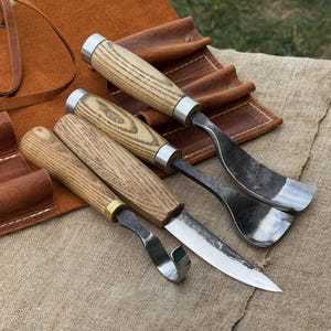 Puede incluir: Cuatro herramientas para tallar madera con mangos de madera y hojas de metal se muestran sobre una superficie beige. Las herramientas incluyen una gubia curva, un cuchillo recto y un cuchillo de gancho. Un rollo de herramientas de cuero marrón está en el fondo.