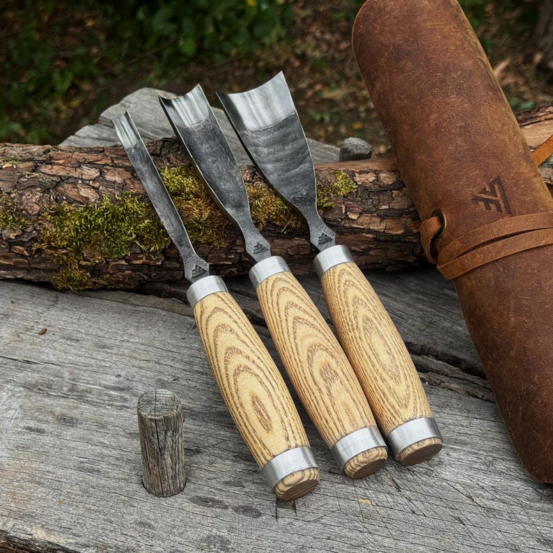 May include: Three wood carving gouges with curved metal blades and light-coloured wood handles featuring prominent grain patterns. Each tool has a silver metal ferrule at the base of the handle and a small logo stamped on the metal neck.