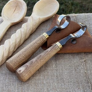 May include: Wooden carving tools and spoons are displayed on a linen surface. Two curved carving knives with wooden handles and brass accents are next to a leather pouch. Two wooden spoons are also visible, one with a twisted handle.
