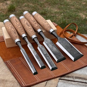 Peut inclure: Un ensemble de cinq ciseaux à bois avec des manches en bois brun clair et des lames en métal sombre et texturé. Les ciseaux sont alignés sur un bloc de bois clair et un étui à outils en cuir brun avec des coutures et des lanières visibles.
