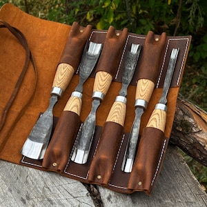 May include: A set of six wood carving chisels with light-colored wooden handles and dark metal blades, organized in a brown leather roll-up tool pouch with white stitching and brass-colored rivets, featuring various blade shapes and sizes.