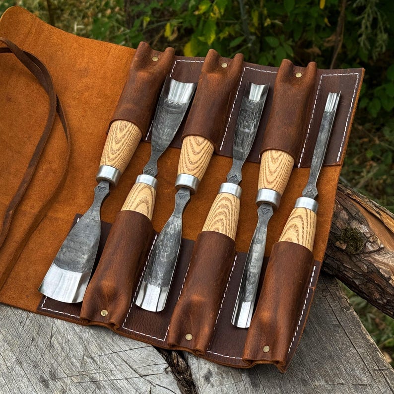 May include: A set of six wood carving chisels with light-colored wooden handles and dark metal blades, organized in a brown leather roll-up tool pouch with white stitching and brass-colored rivets, featuring various blade shapes and sizes.