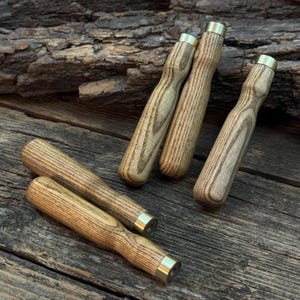 Könnte beinhalten: Eine Sammlung von Holzgriffen mit Messingzwingen. Die Griffe bestehen aus hellfarbenem Holz mit sichtbarer Maserung. Die Messingzwingen befinden sich am Fuß jedes Griffs. Die Griffe sind auf einer Holzoberfläche angeordnet.