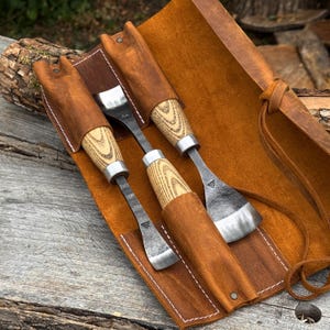 May include: Two metal woodworking tools with light-colored wood handles and silver-colored metal blades, each featuring a small stamped logo. The tools are stored in individual slots within a brown leather roll-up case with white stitching.