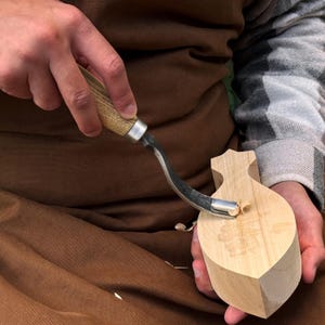 Pu&ograve; includere: Un coltello a uncino in metallo con manico in legno chiaro viene utilizzato per intagliare un pezzo di legno chiaro. L'utensile presenta una lama in acciaio curva che sta rimuovendo un piccolo truciolo di legno dalla superficie del blocco.