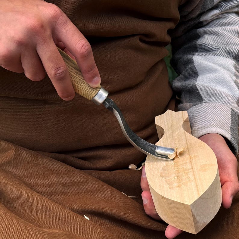 Pu&ograve; includere: Un coltello a uncino in metallo con manico in legno chiaro viene utilizzato per intagliare un pezzo di legno chiaro. L'utensile presenta una lama in acciaio curva che sta rimuovendo un piccolo truciolo di legno dalla superficie del blocco.