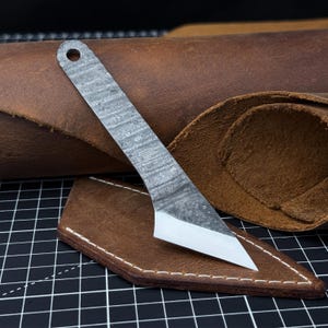 May include: A small, silver-colored knife with a textured pattern and a sharp, angled blade. The knife has a hole at the handle's end. It rests on a brown leather sheath with white stitching, against a backdrop of more brown leather.