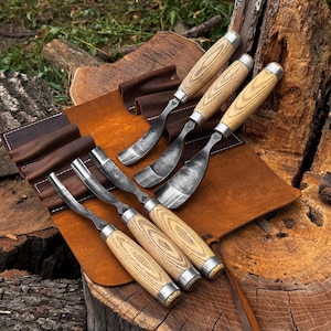 May include: A set of six wood carving gouges with curved metal blades and light-colored wood handles, arranged in a brown leather tool roll. Each gouge features a distinct blade curvature and a metal ferrule connecting the handle to the steel.