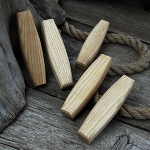 Op de afbeelding: Vijf houten ladeknopen in verschillende tinten lichtbruin en beige zijn gerangschikt op een verweerd houten oppervlak. De knoppen hebben een afgeronde, langwerpige vorm met zichtbare houtnerf. Een stuk touw is zichtbaar op de achtergrond.