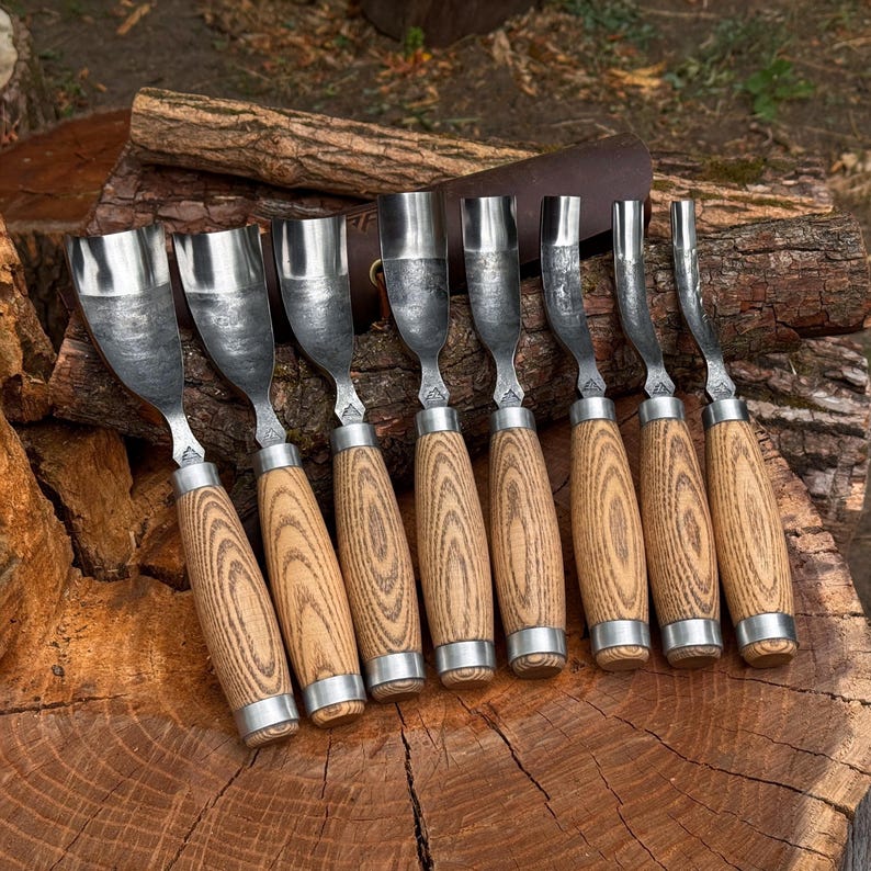 May include: A set of eight wood carving gouges with curved metal blades and light brown wooden handles. Each tool features a polished steel head with a distinct curvature, securely attached to a textured wood grip with a silver metal ferrule.