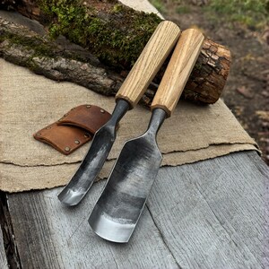 May include: Two woodworking gouges with wooden handles and metal blades, resting on a wooden surface. A small, brown leather sheath is also present. The tools are designed for carving and shaping wood.