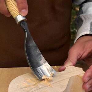Pu&ograve; includere: Una sgorbia curva per intaglio del legno con una lama in metallo lucido e un manico in legno chiaro. L'utensile presenta un marchio distintivo del produttore vicino alla ghiera e rimuove un sottile truciolo di legno chiaro da una superficie.